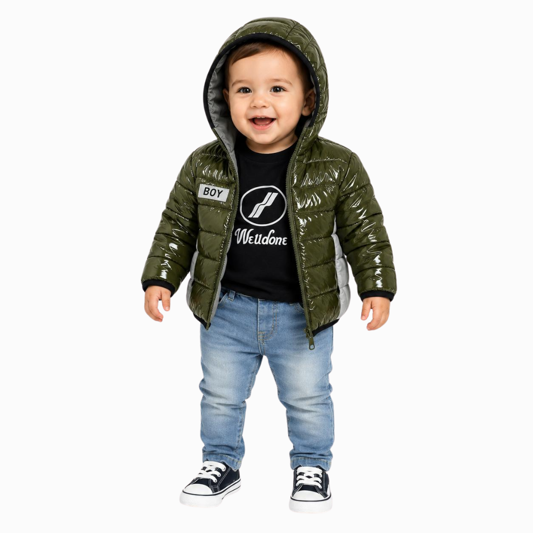 A toddler stands against a light gray background wearing a shiny olive green hooded puffer jacket over a black graphic T-shirt, blue jeans, and black-and-white sneakers.