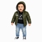 A toddler stands against a light gray background wearing a shiny olive green hooded puffer jacket over a black graphic T-shirt, blue jeans, and black-and-white sneakers.