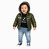 A toddler stands against a light gray background wearing a shiny olive green hooded puffer jacket over a black graphic T-shirt, blue jeans, and black-and-white sneakers.
