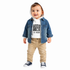 A toddler standing against a white background wearing a blue denim jacket over a white graphic hoodie, tan pants, and navy sneakers.
