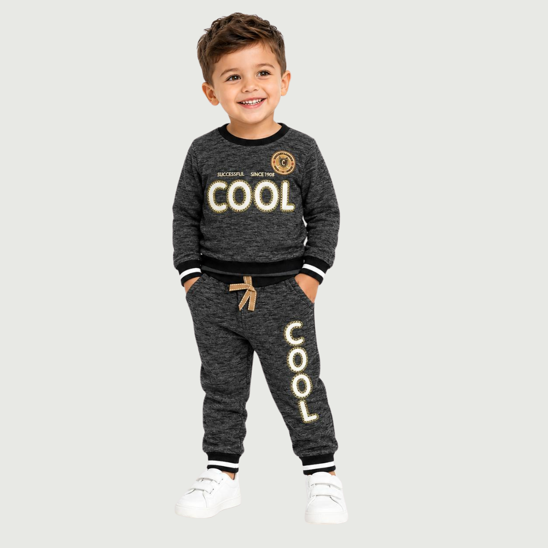 A young boy smiling and wearing a dark gray heathered two-piece sweatshirt and jogger pants set with gold and white 'COOL' graphics, standing against a light gray background.