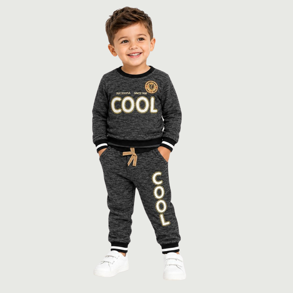 A young boy smiling and wearing a dark gray heathered two-piece sweatshirt and jogger pants set with gold and white 'COOL' graphics, standing against a light gray background.