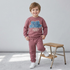 A young child wearing a dusty rose sweatshirt and matching sweatpants set with a blue graphic on the chest, standing next to a small wooden step stool in a bright room.