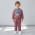 A young child wearing a dusty rose sweatshirt and matching sweatpants set with a blue graphic on the chest, standing against a light gray wall.