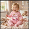 Baby in a pink 'The Princess has arrived' onesie sitting on a bed with teddy bears and toys.