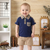 Child wearing a navy blue shirt with plaid accents and beige shorts in a home setting.