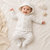 A baby lying on a cream-colored blanket, wearing a matching hooded knit romper set in off-white with a subtle light beige pattern. The set includes a long-sleeve top with buttons and matching footed pants.