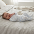 A baby lying on a light beige linen bed, wearing a white footed sleeper with light blue heart patterns. A small plush toy is tucked near the baby's head.