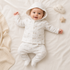 A baby lying on a cream-colored blanket, wearing a matching hooded knit romper set in off-white with a subtle light beige pattern. The set includes a long-sleeve top with buttons and matching footed pants.