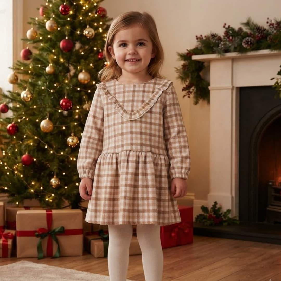 A young girl stands in a cozy holiday living room wearing a beige and brown gingham dress with a ruffled collar, white tights, and smiling. A decorated Christmas tree with red and gold ornaments, wrapped gifts, and a fireplace with green garlands are visible in the background.