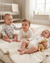 Three babies sitting on a bed in a nursery with a crib and window in the background.