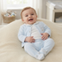 A smiling baby sitting on a cream-colored textured blanket in a bright nursery, wearing a light blue footed sleeper with a white inner top and matching booties.