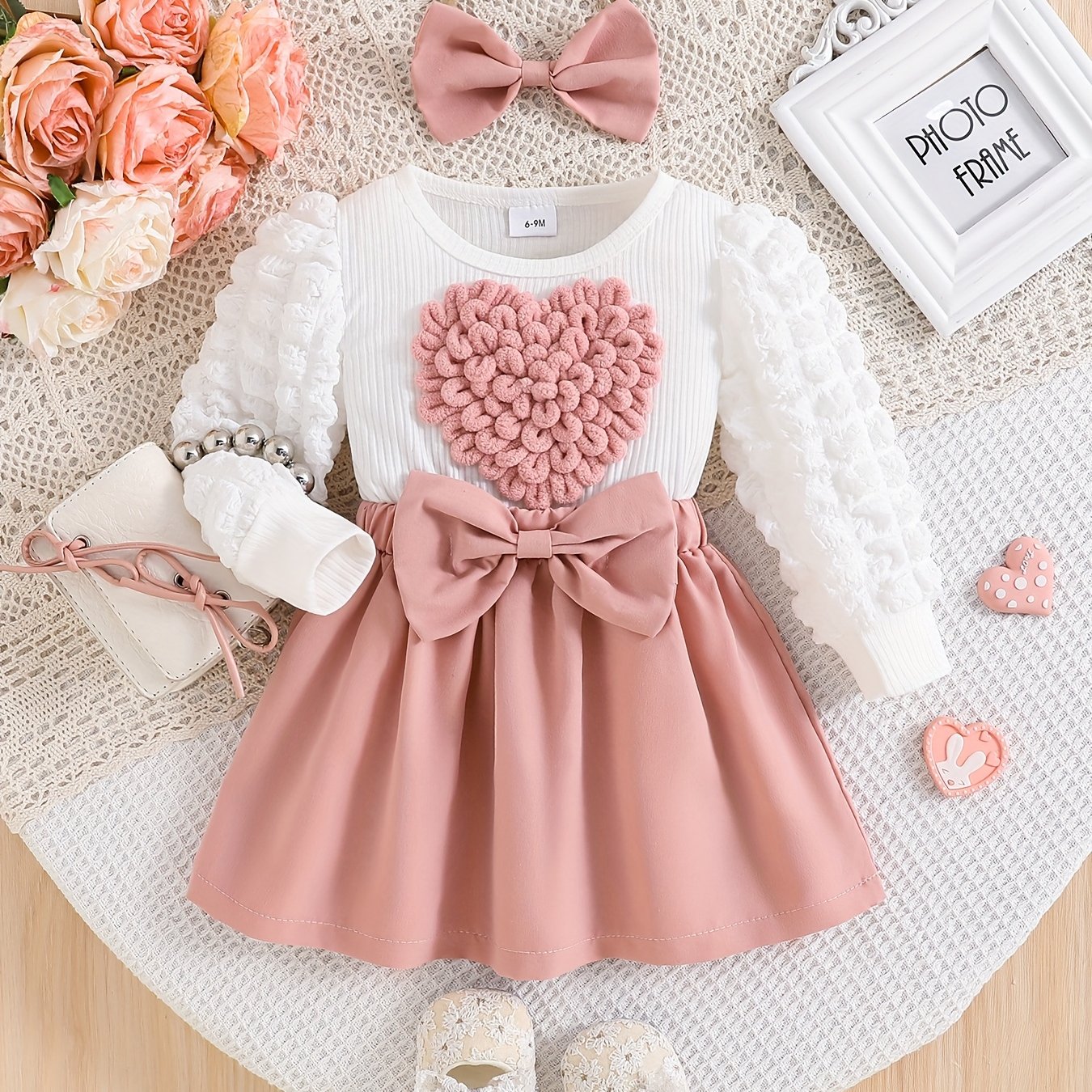 Children's dress with pink heart design and bow, surrounded by flowers and a photo frame on a textured surface.