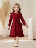 Young girl wearing a red dress with long sleeves and a belt, standing in a room with a white chair and decorative plant.