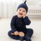 Baby in navy outfit sitting on a light-colored floor with a white crib in the background