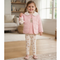 A young girl stands in a bright living room wearing a light pink quilted jacket with a white faux-fur collar, a matching pink top, and cream leggings with a teddy bear pattern. She is holding a small plush white bear.