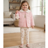 A young girl stands in a bright living room wearing a light pink quilted jacket with a white faux-fur collar, a matching pink top, and cream leggings with a teddy bear pattern. She is holding a small plush white bear.